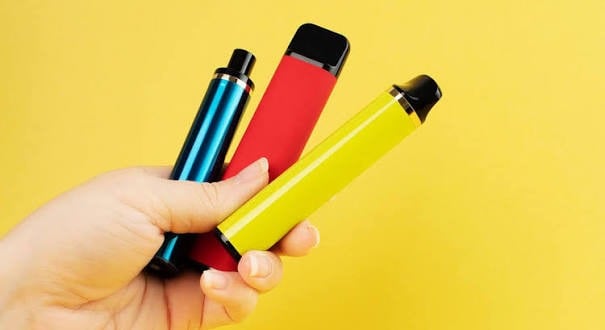 Hand holding three colourful vape devices against a yellow background